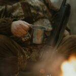 Une personne en tenue de camouflage profite d'un feu de camp avec une tasse en fer-blanc à l'extérieur.