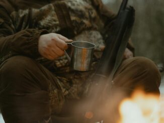 Une personne en tenue de camouflage profite d'un feu de camp avec une tasse en fer-blanc à l'extérieur.
